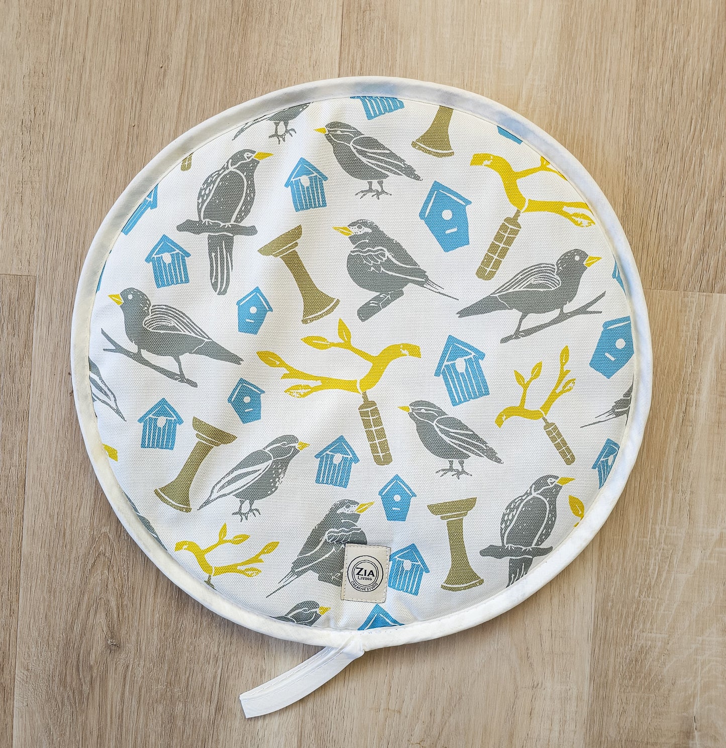 Bird House print round aga hob cover by ZIA LIVING, featuring illustrated birds, birdhouses and branches in soft grey, blue and yellow tones, shown on a wooden surface.