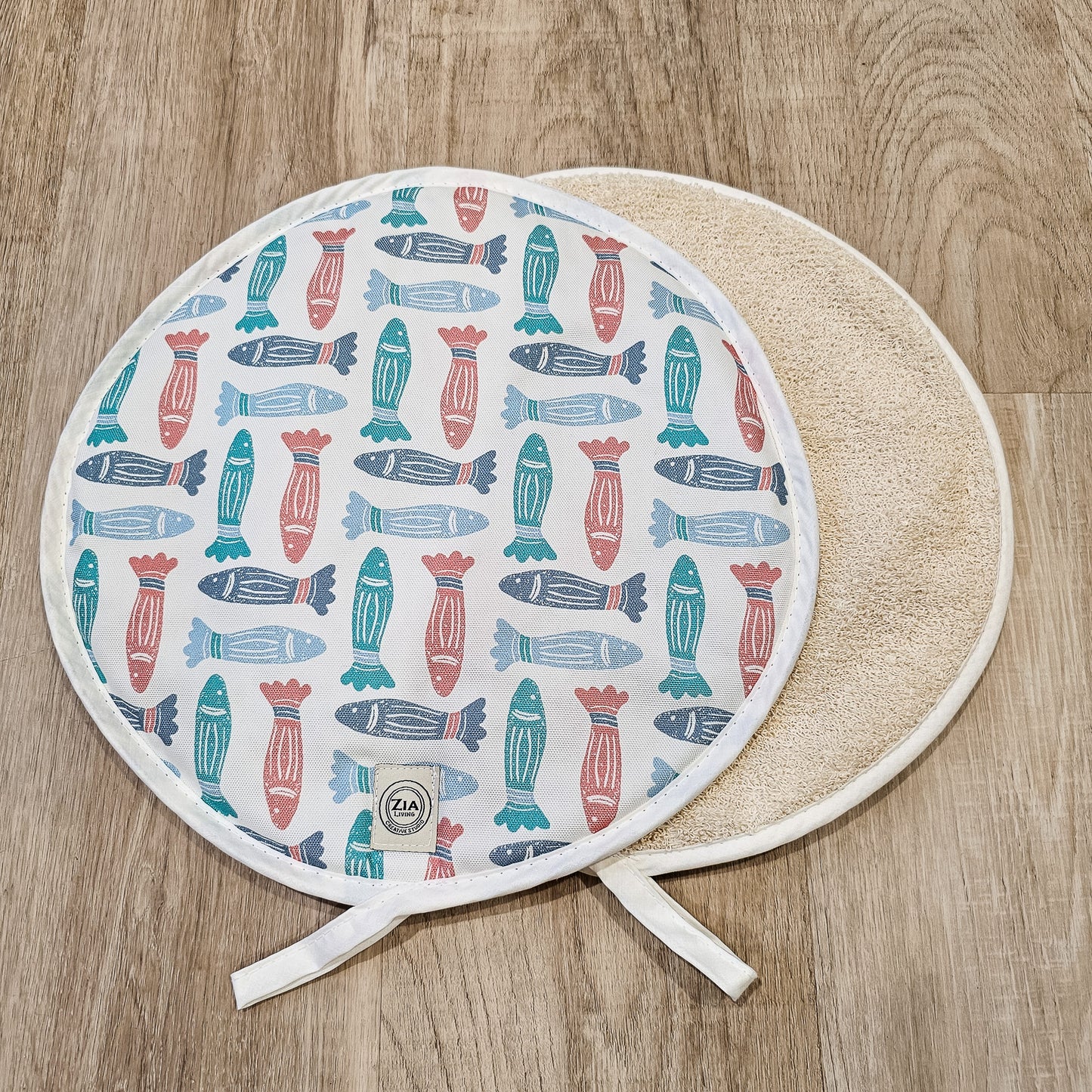 Fish print round kitchen cover by ZIA LIVING, featuring a repeating red, teal and light‑blue fish motif on cotton‑linen, shown with a natural textured underside on a wooden surface.