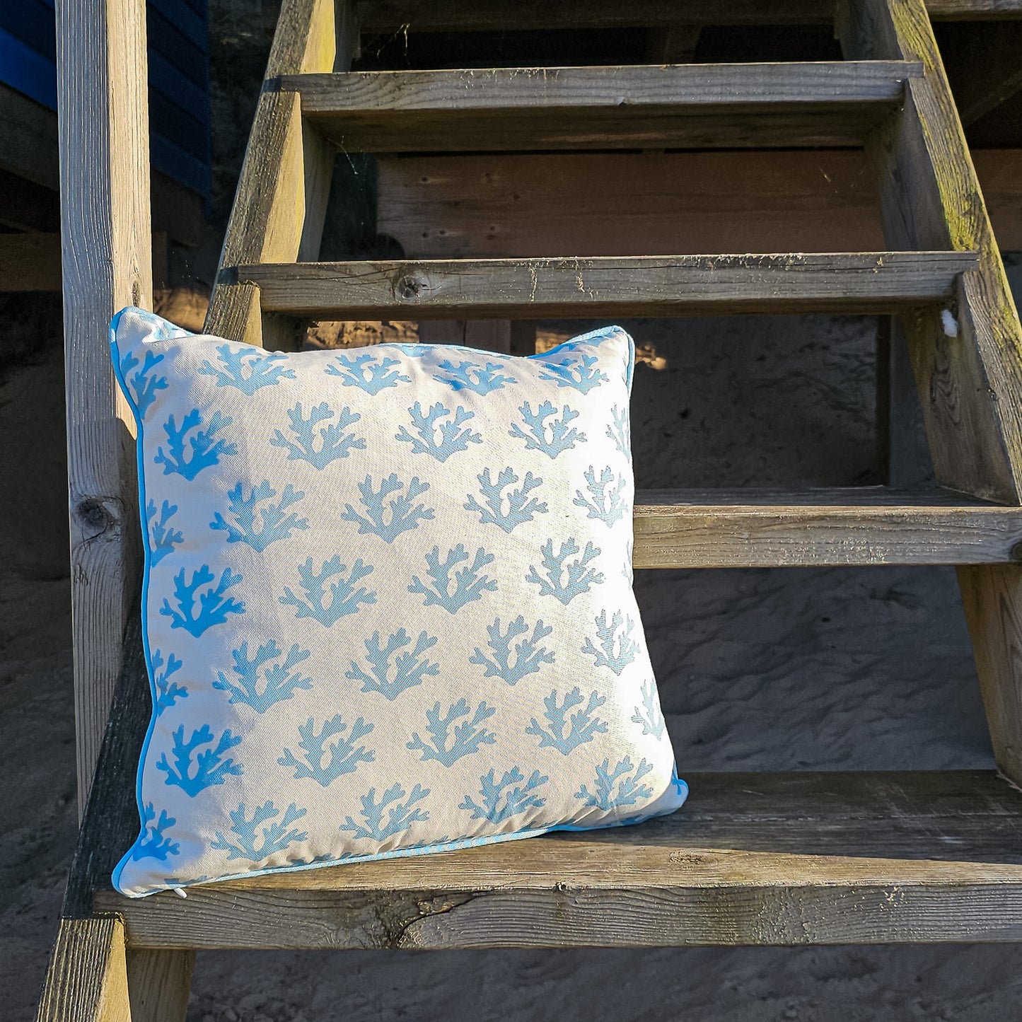 Sky blue seaweed print cushion by ZIA LIVING, featuring a white and blue coral motif with blue piping, styled on a wooden steps