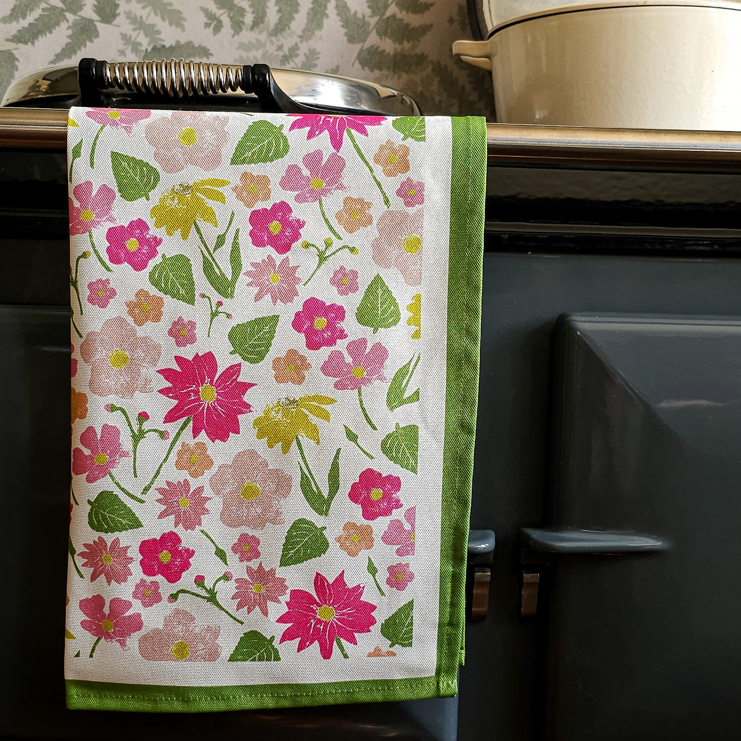 Flower Garden tea towel hanging over a aga rail, featuring a vibrant mix of pink, yellow and orange florals with green leaves and a bold green border — ZIA LIVING.