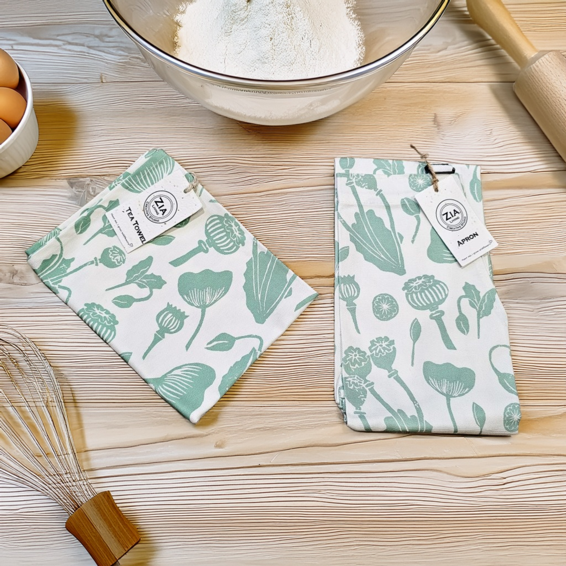ZIA LIVING apron and tea towel set with green botanical print displayed on a wooden kitchen surface beside baking ingredients, eggs, and utensils