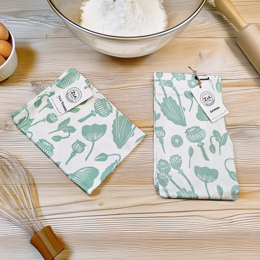 ZIA LIVING apron and tea towel set with green botanical print displayed on a wooden kitchen surface beside baking ingredients, eggs, and utensils