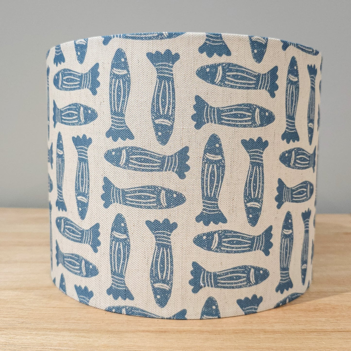 Blue Fishes drum lampshade featuring simple hand‑drawn fish motifs on a warm neutral background, styled on a wooden surface — ZIA LIVING.