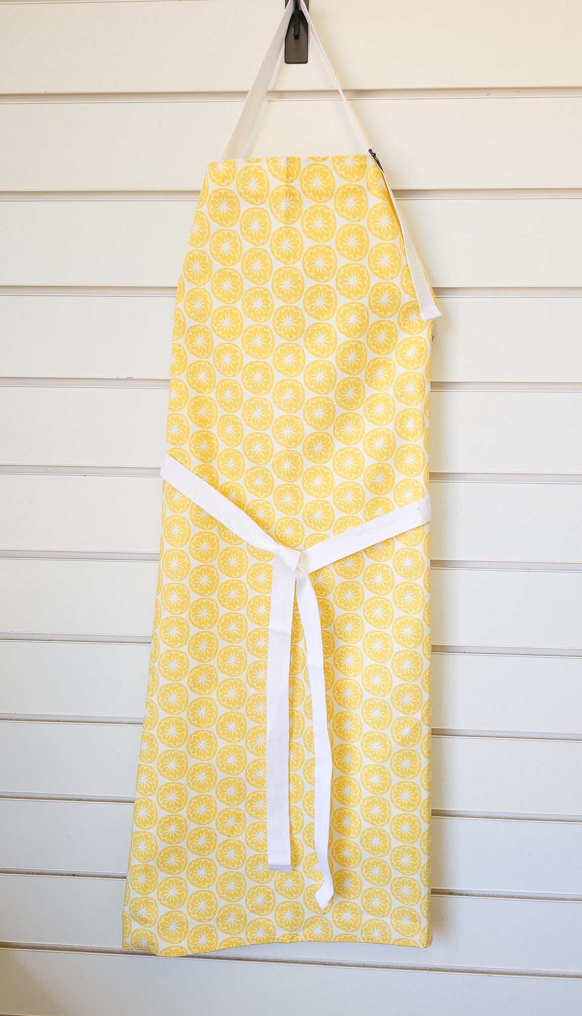 A yellow Poppy Seed Head apron from ZIA LIVING hanging with wooden background