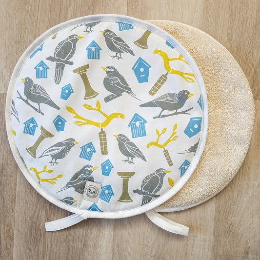 A Bird House print round aga hob cover from ZIA LIVING laid on a wooden surface, showing the bird and birdhouse pattern.