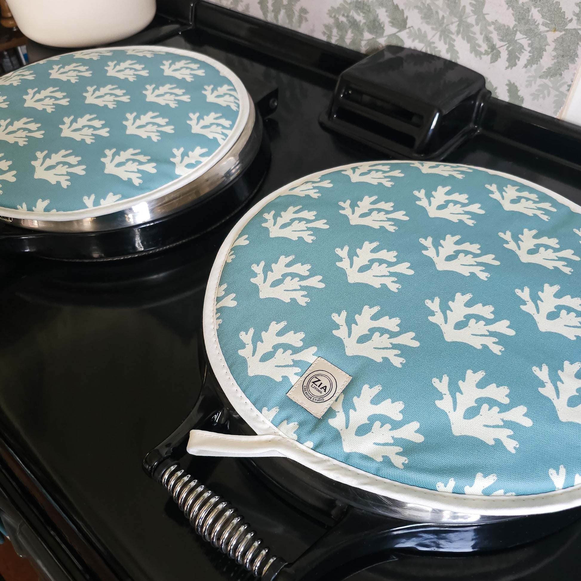 Blue seaweed print Aga and hob covers by ZIA LIVING, featuring a white coral motif on a deep‑blue cotton base, shown on a glossy black stove with metal handles.