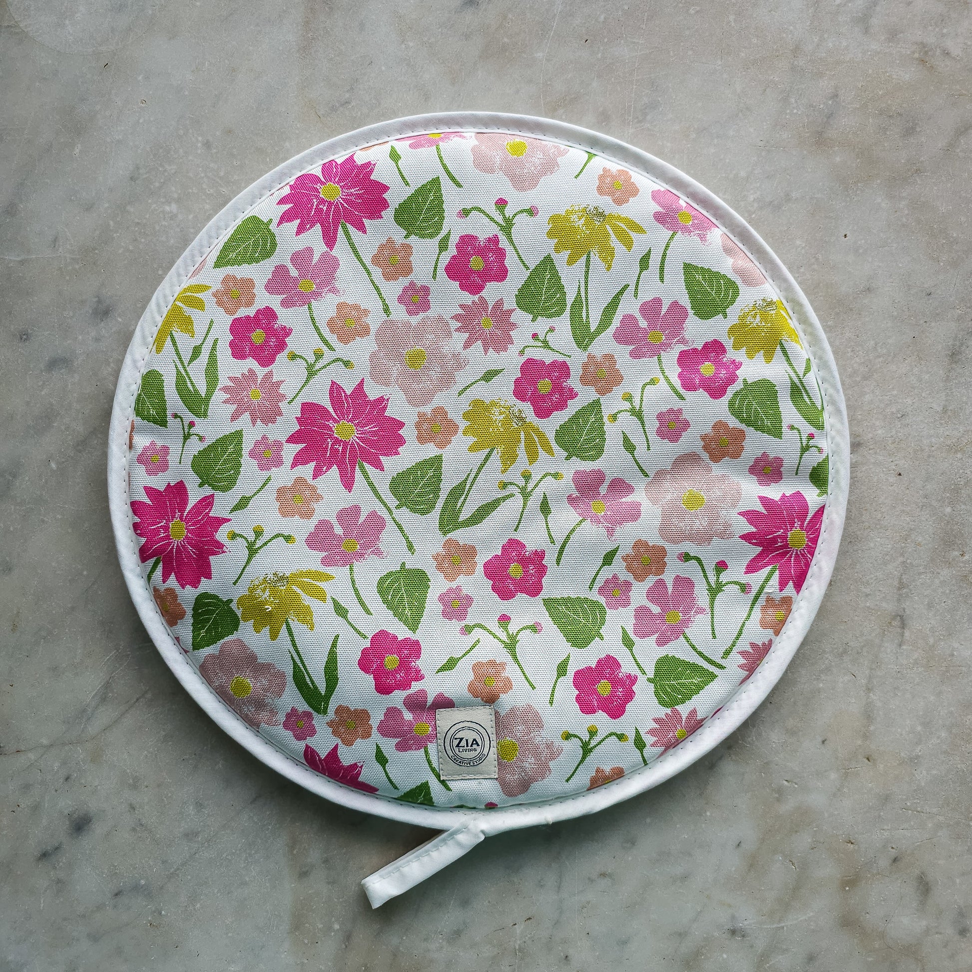 Floral quilted round hob cover by ZIA LIVING, featuring pink, yellow and orange flowers with green leaves on a white background, shown on a light stone surface with a stitched border and hanging loop.