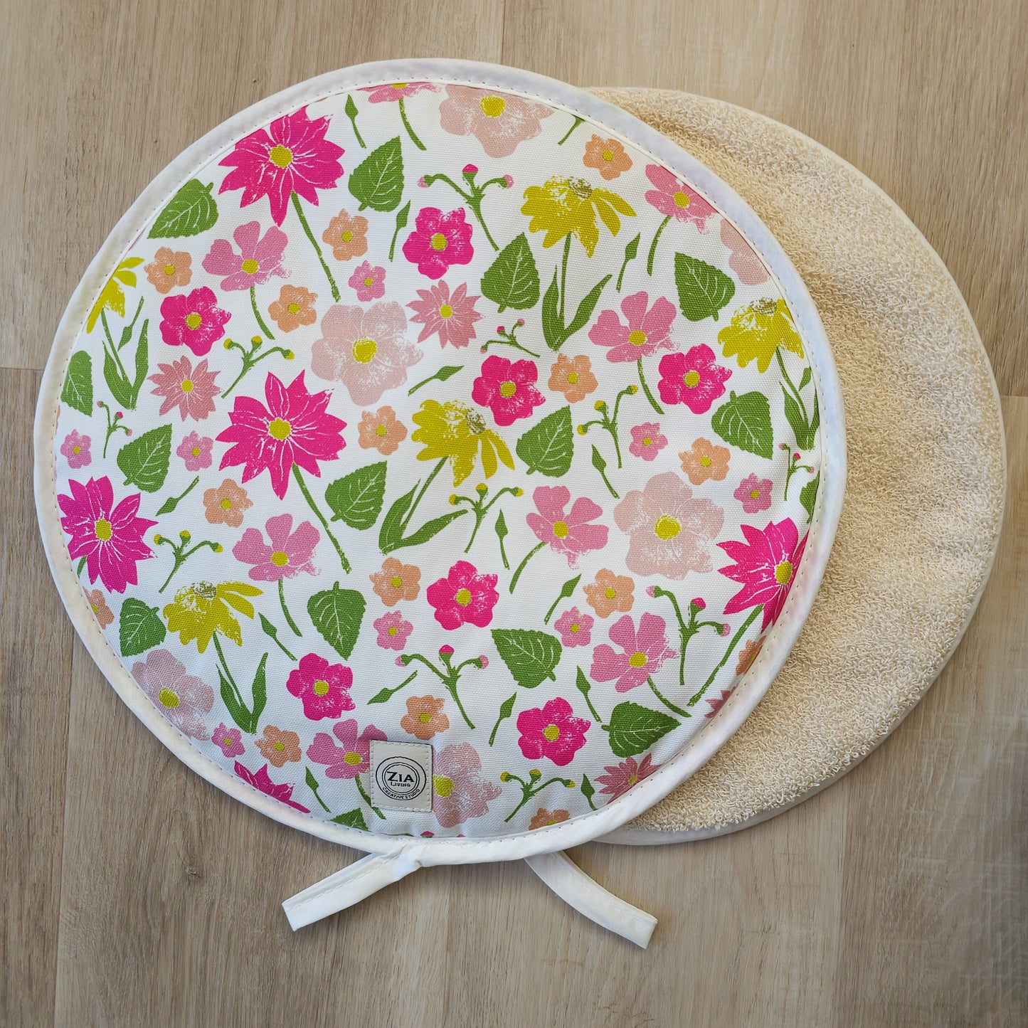 Floral quilted round hob cover by ZIA LIVING, featuring pink, yellow and orange flowers with green leaves on a white background, shown on a light wooden surface with a stitched border, towelling reverse and hanging loop.