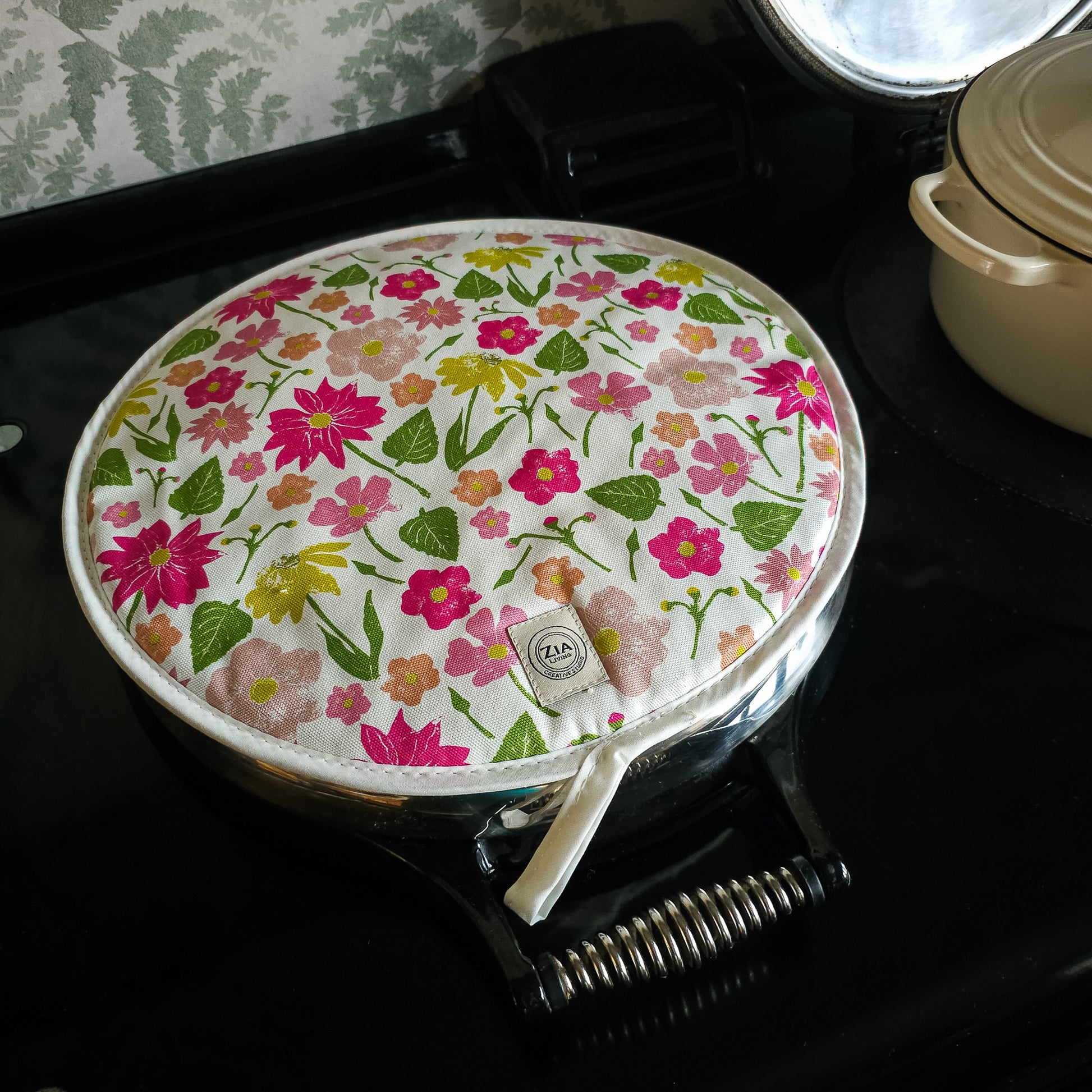 Floral quilted round hob cover by ZIA LIVING, featuring pink, yellow and orange flowers with green leaves on a white background, shown on a aga top with a stitched border and hanging loop.