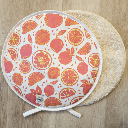Citrus print round hob cover by ZIA LIVING, featuring orange and grapefruit motifs on a white cotton base, shown with a natural textured underside on a wooden surface.