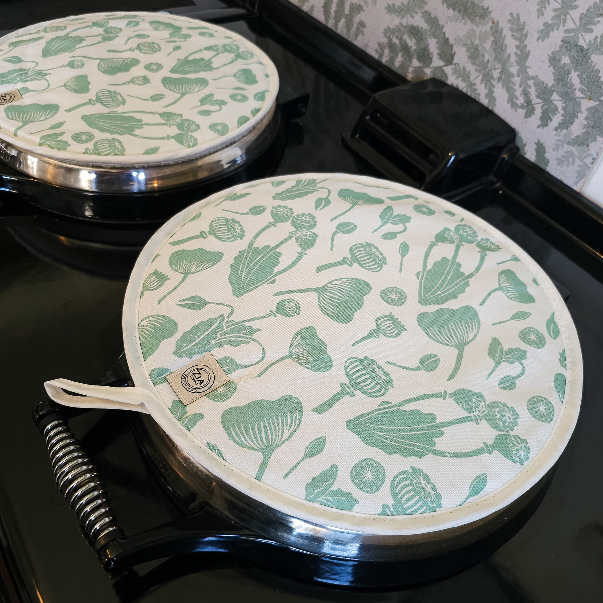 Green botanical print Aga and hob covers by ZIA LIVING, featuring a cream cotton base with stylised green floral motifs, shown on a glossy black Aga stove with metal handles.