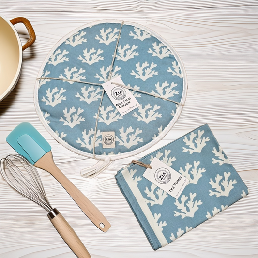A round Seaweed print aga hob cover and matching tea towel from ZIA LIVING arranged on a white wooden surface alongside kitchen utensils and a cream pot.