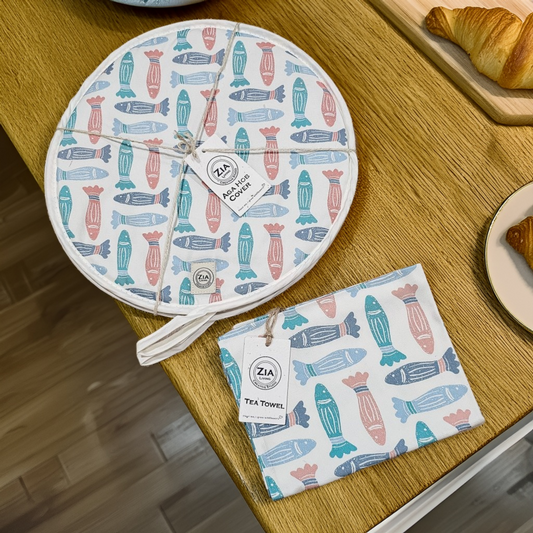 A round Fishes print aga hob cover and matching tea towel from ZIA LIVING arranged on a wooden worktop with Danish pastries.
