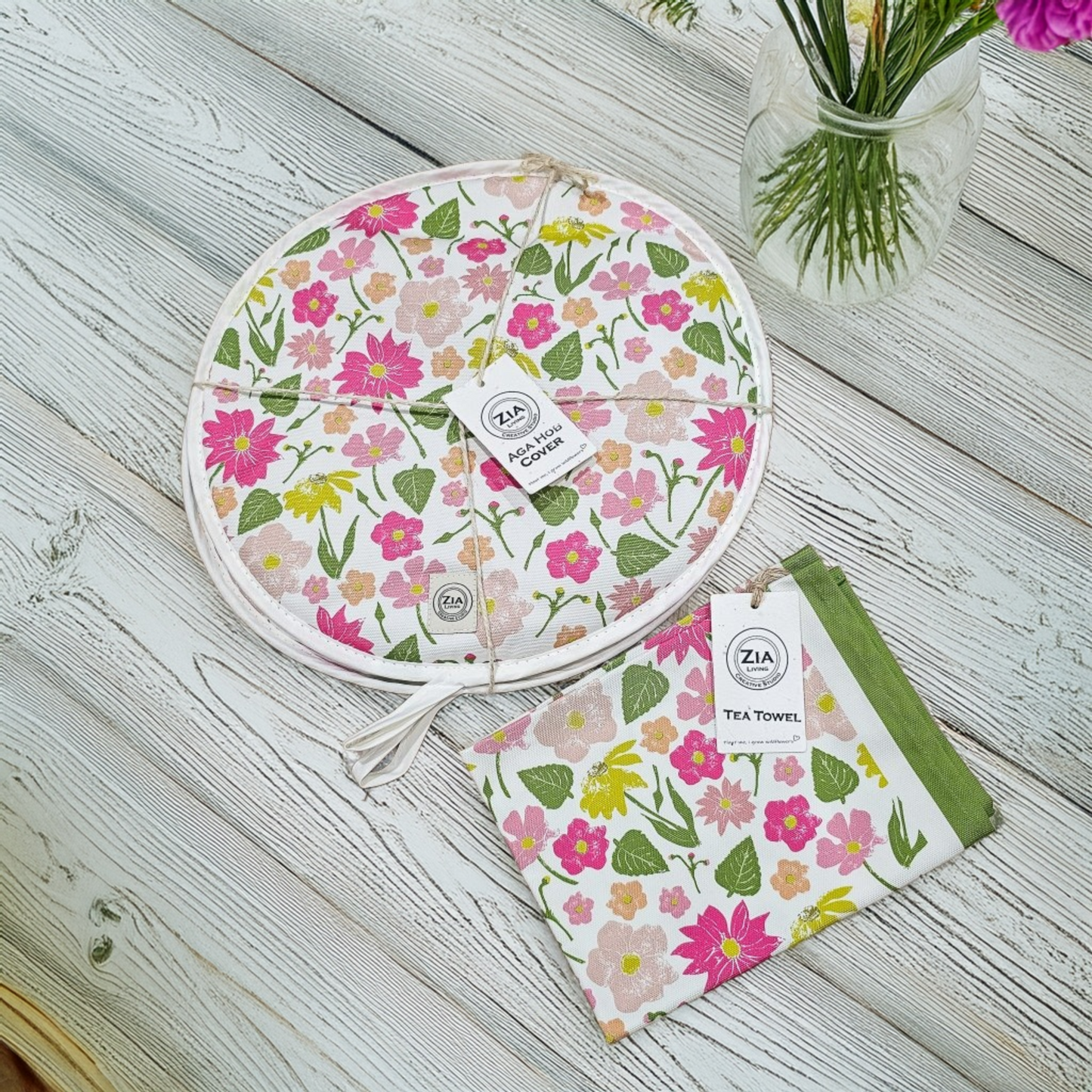 A Flower Garden print round aga hob cover and matching tea towel from ZIA LIVING arranged on a white wooden surface beside a small jar of flowers.