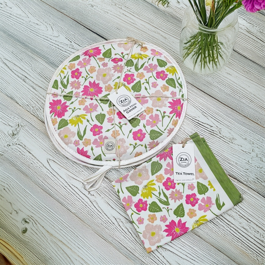 A Flower Garden print round aga hob cover and matching tea towel from ZIA LIVING arranged on a white wooden surface beside a small jar of flowers.