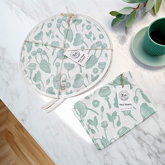A Green Botanicals print round aga hob cover and matching tea towel from ZIA LIVING arranged on a marble surface beside a green cup and  plant leaf