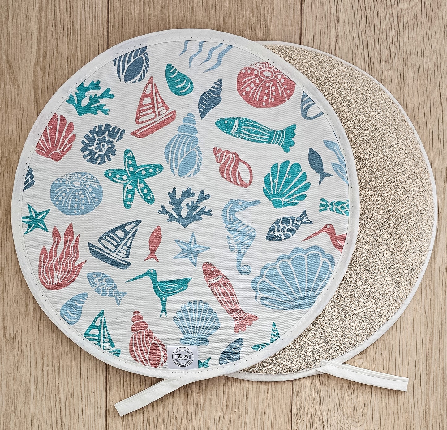 Under the Sea round Aga and hob cover by ZIA LIVING, featuring a colourful coastal lino‑print pattern with sea creatures, shells and boats on cotton, shown with a natural textured underside on a wooden surface.