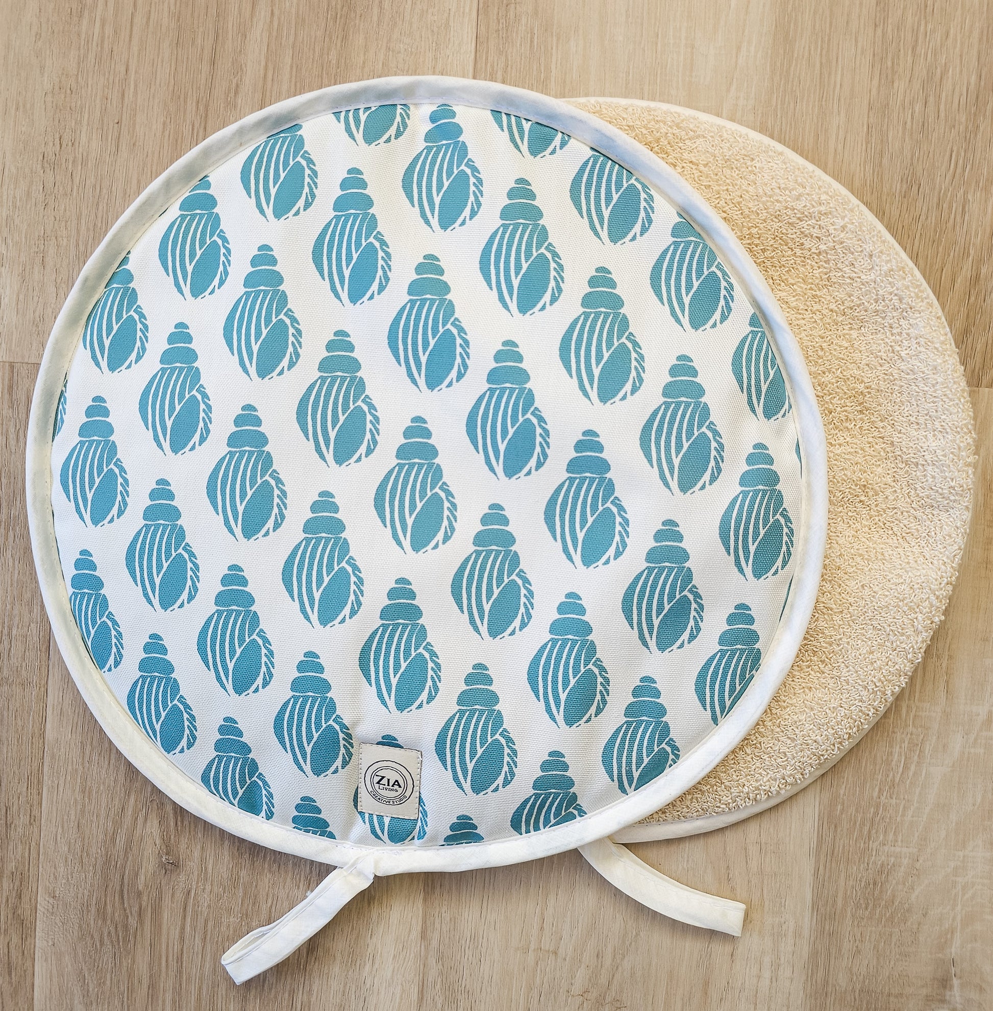 Sea Blue Shell round Aga and hob cover by ZIA LIVING, featuring a blue shell lino‑print pattern on cotton with fabric ties, shown with a plain natural underside on a wooden surface.
