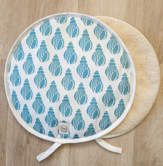 Sea Blue Shell round Aga and hob cover by ZIA LIVING, featuring a blue shell lino‑print pattern on cotton with fabric ties, shown with a plain natural underside on a wooden surface.