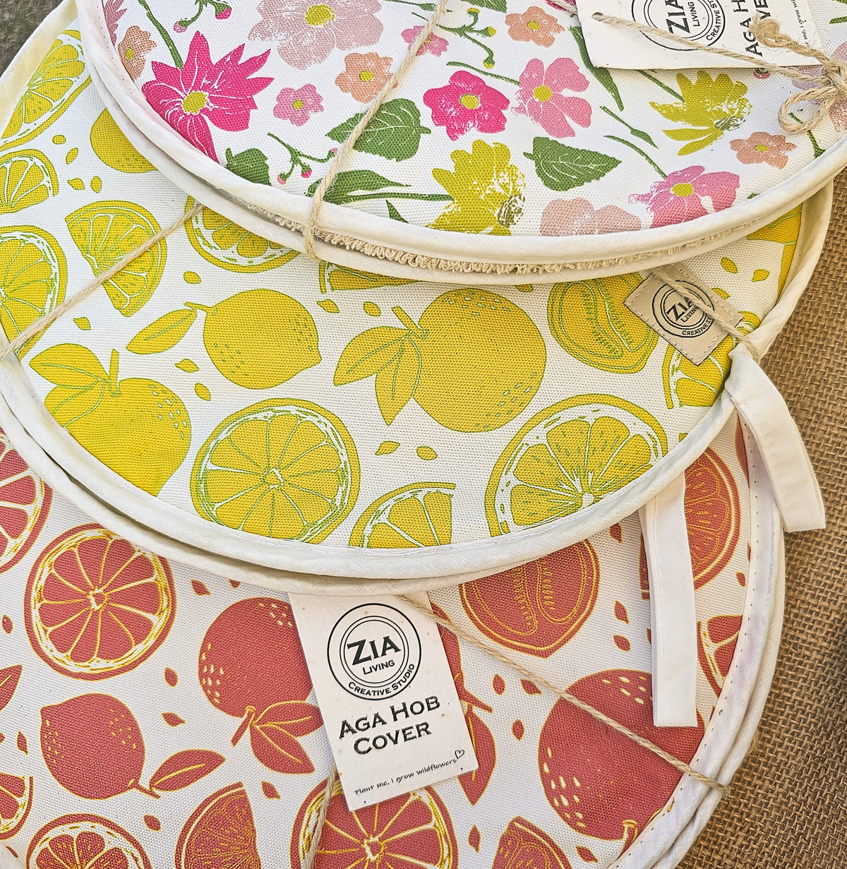 Three round woven aga hob covers kitchen stacked together on a hessian surface. The top cover has a pink and green floral print, the middle cover has a yellow lemon and orange citrus print, and the bottom cover has a red and orange citrus print.