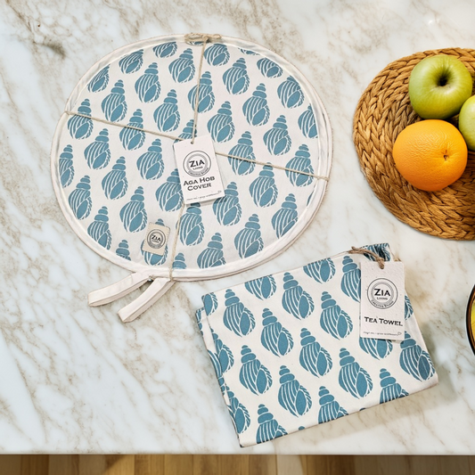 A round Shell print aga hob cover and matching tea towel from ZIA LIVING arranged on a marble worktop beside a woven placemat with fruit