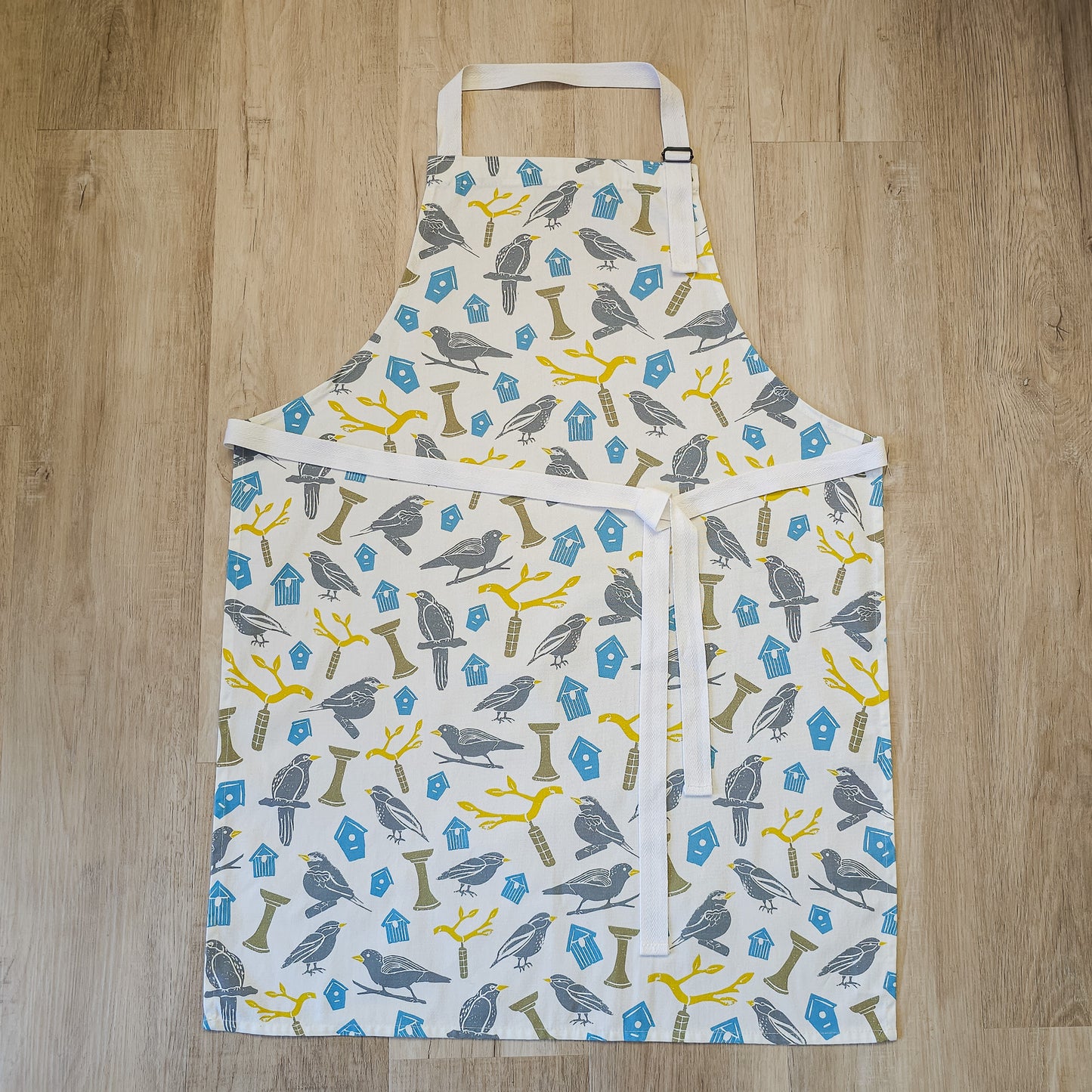 Birdhouse print cotton apron by ZIA LIVING, featuring illustrated birds, feeders and branches in soft grey, yellow and blue tones, shown flat on a wooden surface with white neck and waist ties.