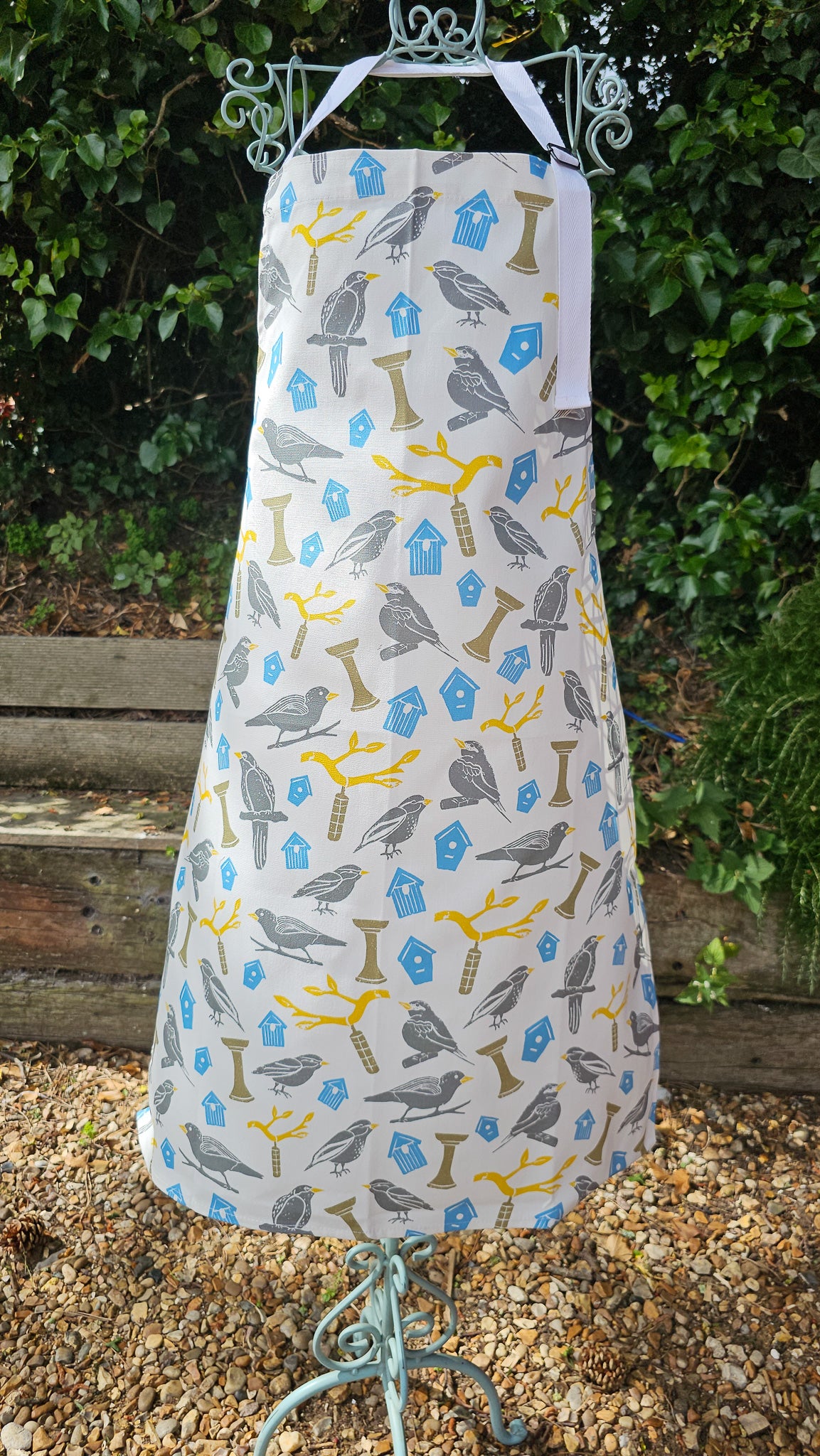 Birdhouse print cotton apron by ZIA LIVING, featuring illustrated birds, feeders and branches in soft grey, yellow and blue tones, shown on a display in garden