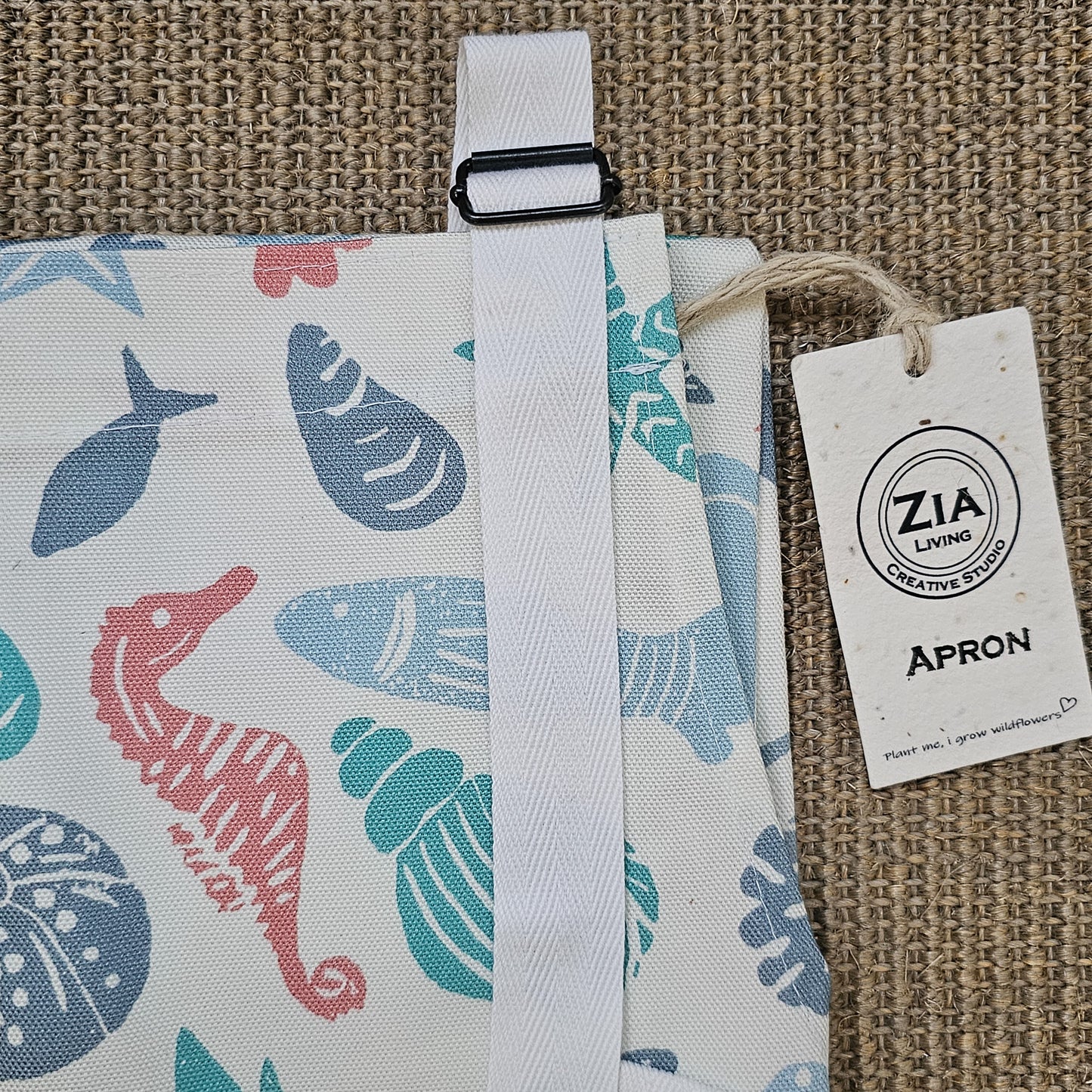 Under the Sea print cotton apron by ZIA LIVING, featuring colourful coastal illustrations of shells, starfish, coral and sea creatures on a white background, folded showing buckle
