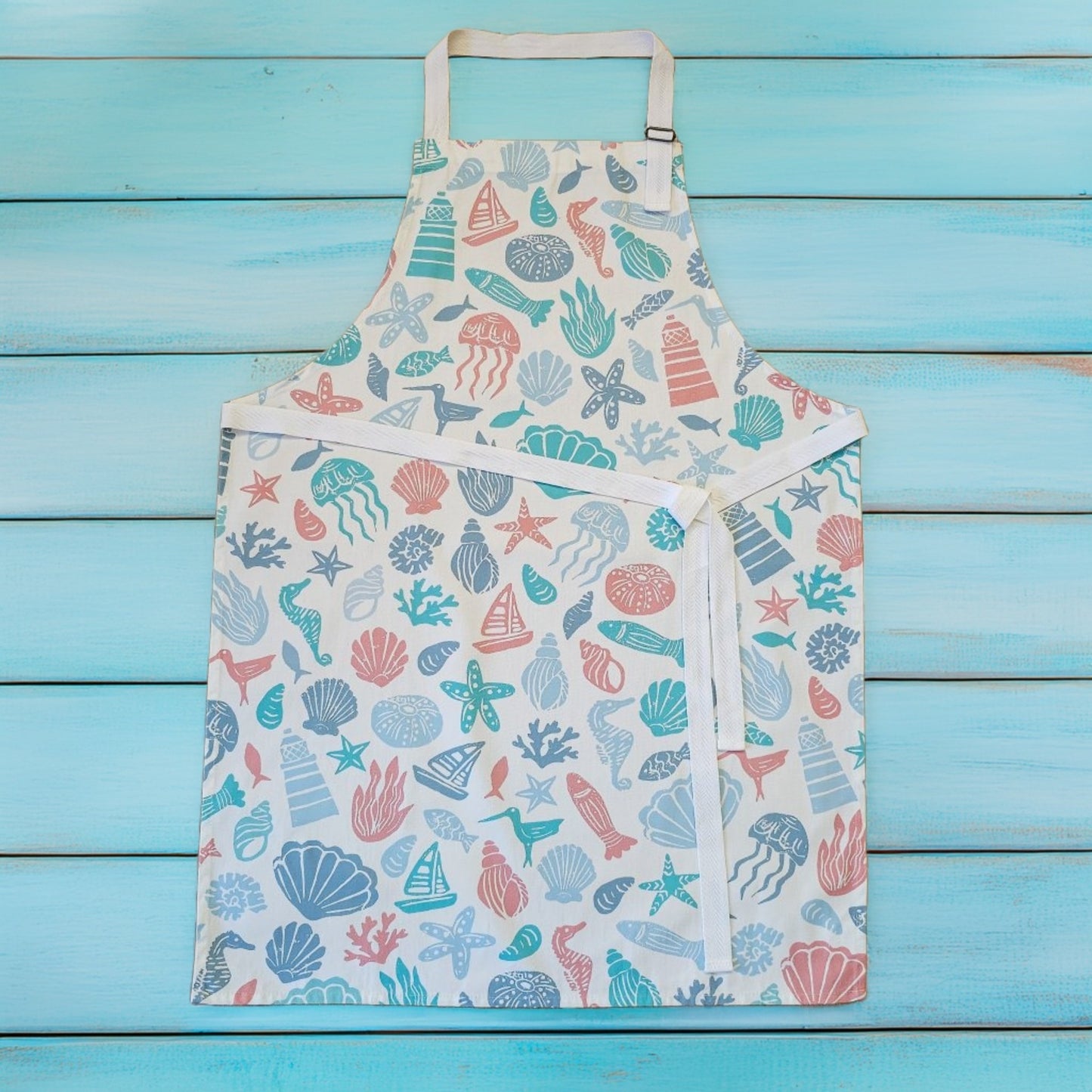 Under the Sea print cotton apron by ZIA LIVING, featuring colourful coastal illustrations of shells, starfish, coral and sea creatures on a white background, shown flat on a turquoise wooden surface with white neck and waist ties.