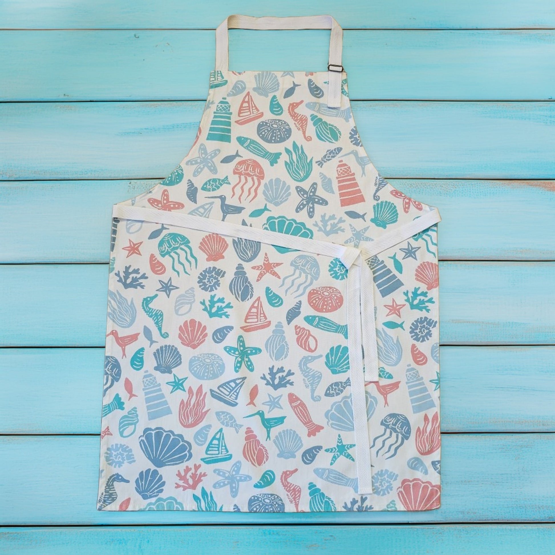 Under the Sea print cotton apron by ZIA LIVING, featuring colourful coastal illustrations of shells, starfish, coral and sea creatures on a white background, shown flat on a turquoise wooden surface with white neck and waist ties.