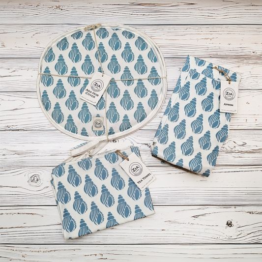 A complete blue Shell kitchen set from ZIA LIVING—two aga hob covers, an apron, and a tea towel—arranged together on a wooden surface to show this coastal pattern.