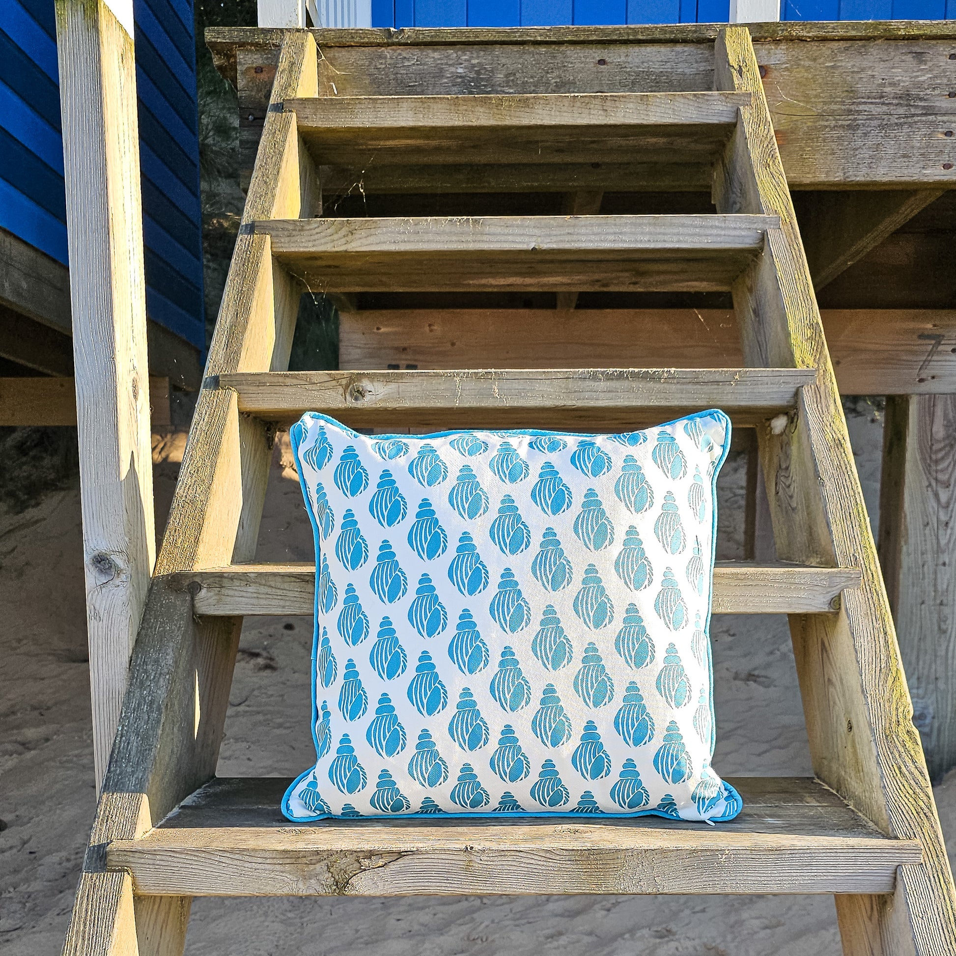 Sea Blue Shell cushion featuring a repeating hand‑carved shell motif in blue on a white background, photographed on a wooden steps — ZIA LIVING.