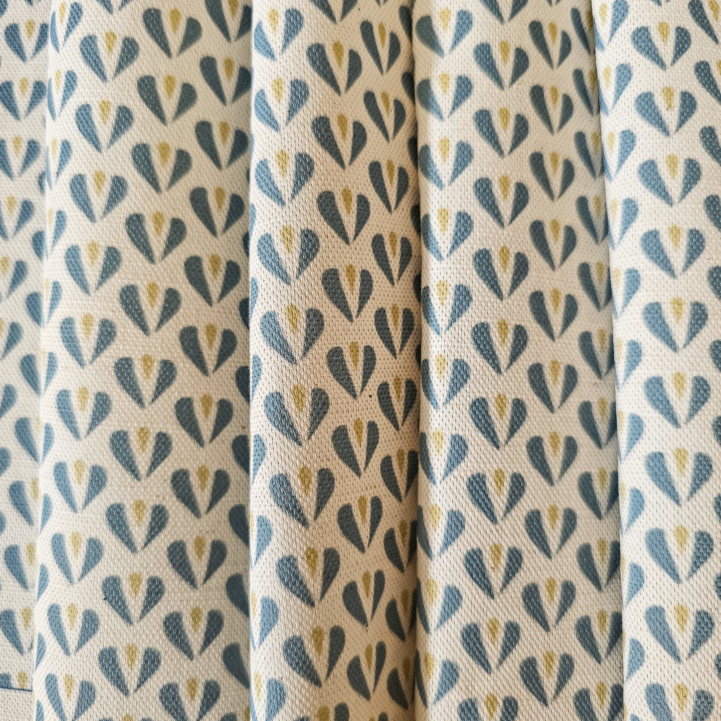 Carina, heart shaped motif in blue and sand colours on neutral fabric, shown as drapes, by Zia Living
