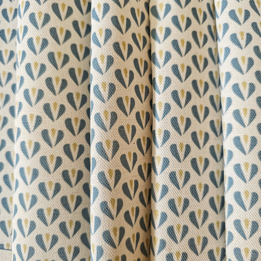Carina, heart shaped motif in blue and sand colours on neutral fabric, shown as drapes, by Zia Living