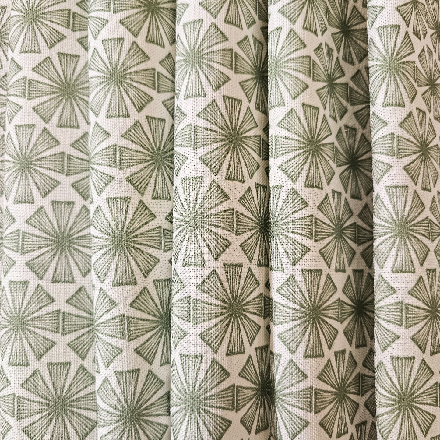 Stella cotton linen fabric featuring a bold green radiating motif in a repeating geometric pattern on an off‑white background. shown as draped fabric. ZIA LIVING.