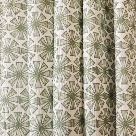Stella cotton linen fabric featuring a bold green radiating motif in a repeating geometric pattern on an off‑white background. shown as draped fabric. ZIA LIVING.