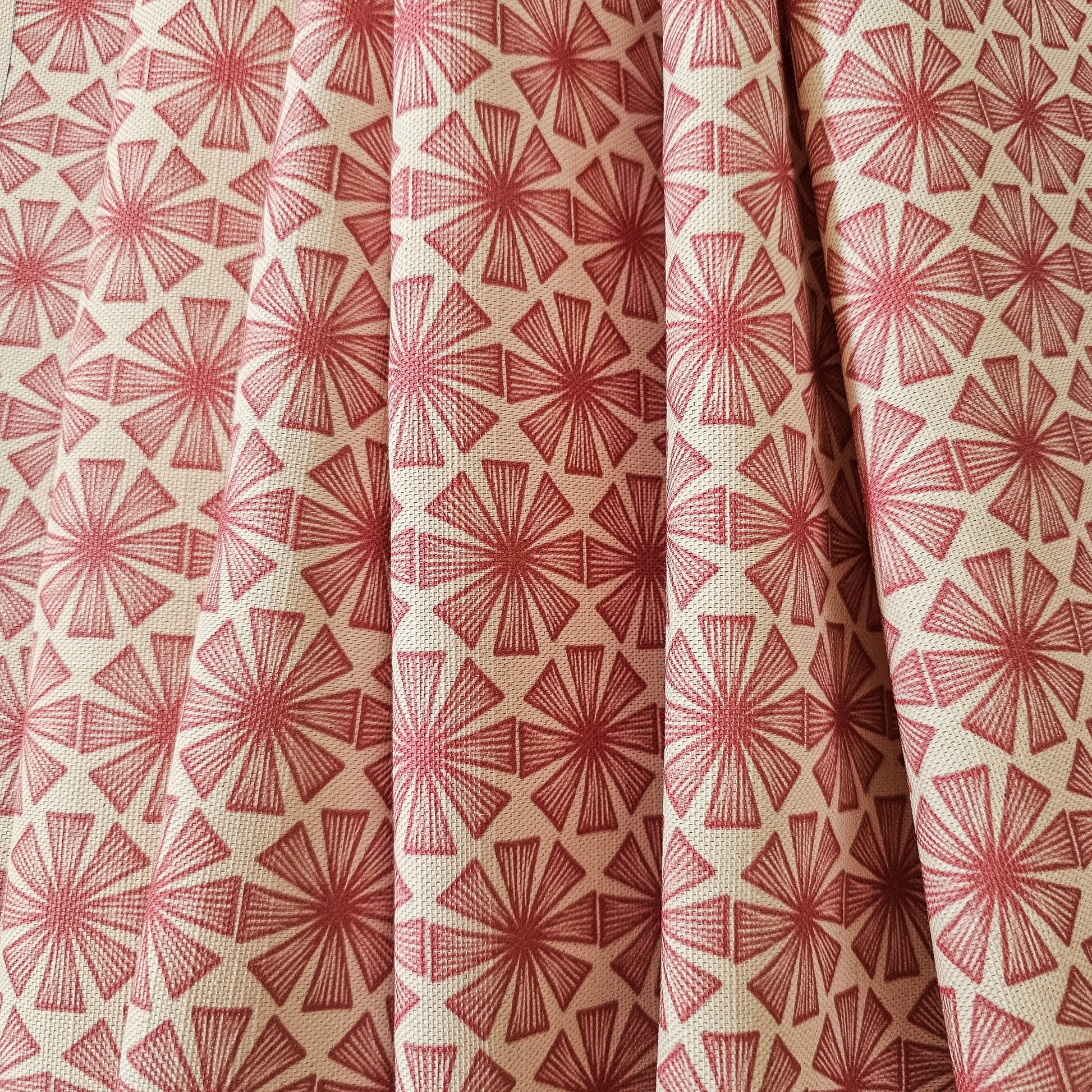 Stella cotton linen fabric featuring a bold red radiating motif in a repeating geometric pattern on an off‑white background. shown as draped fabric. ZIA LIVING.