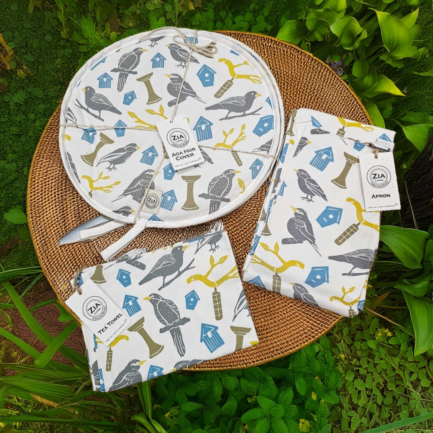 A complete Bird House kitchen linen set from ZIA LIVING—apron, tea towel, and aga cover—styled together on a woven mat in a garden setting