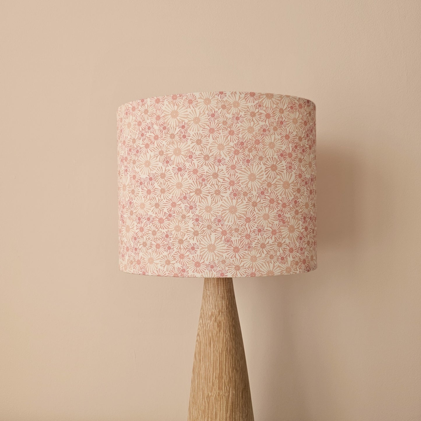 An oval lampshade in a pink Daisy print floral print fabric displayed on a smooth wooden lamp base against a neutral background.