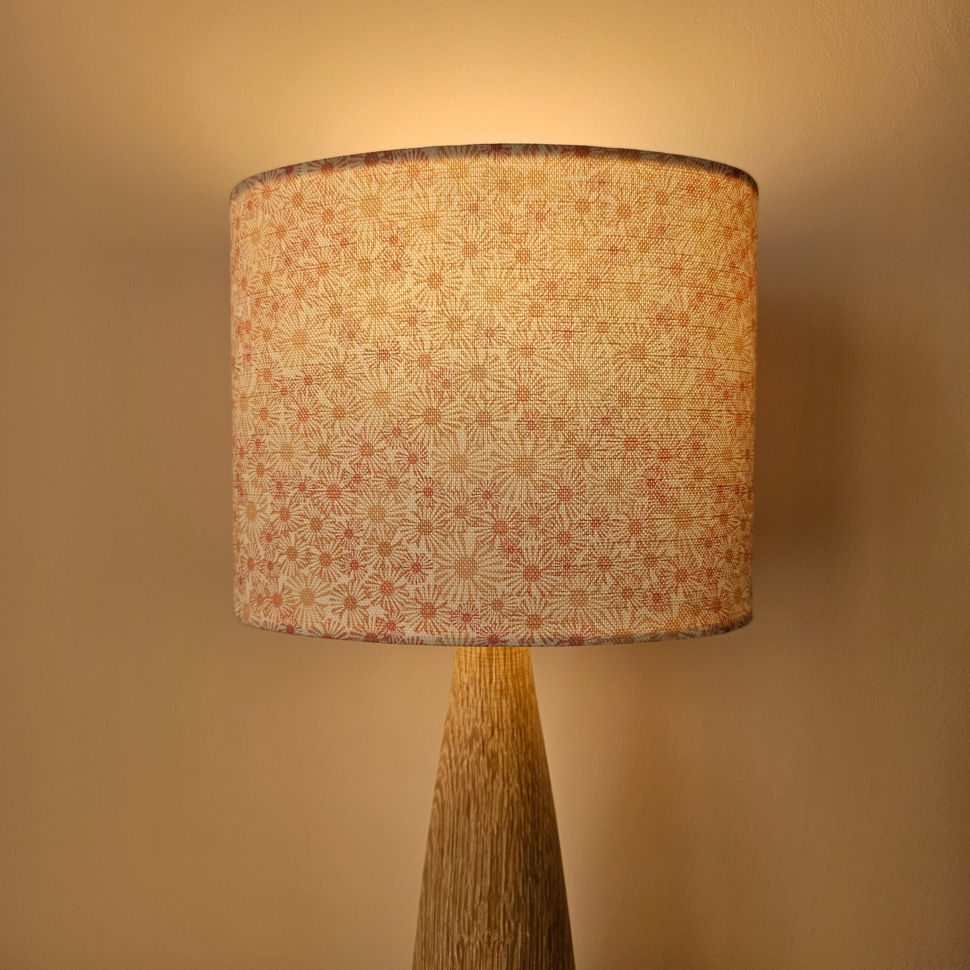 An oval lampshade in a pink Daisy print floral print fabric displayed on a smooth wooden lamp base, with soft glow from light bulb against a neutral background.
