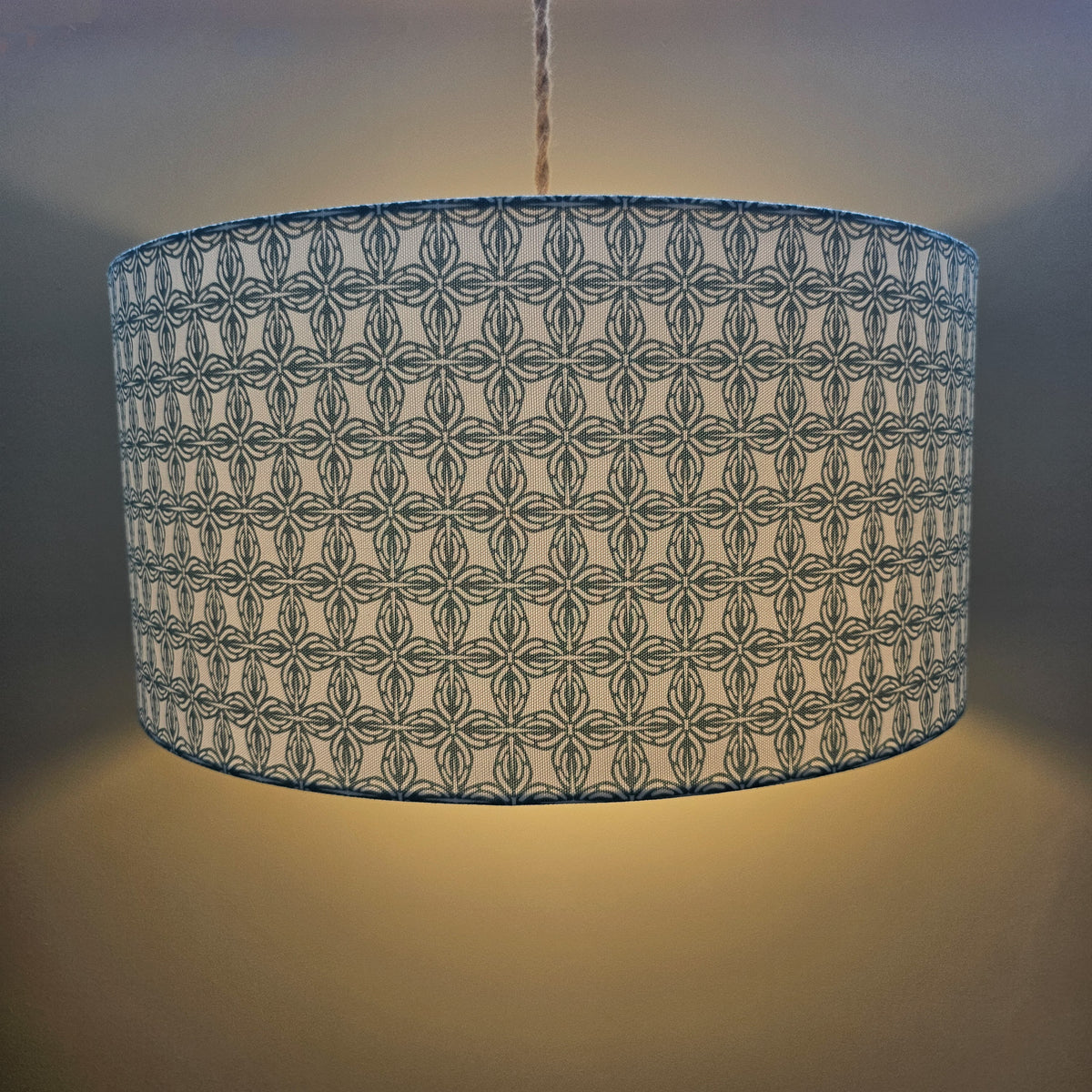 A drum-shaped pendant lampshade with a repeating blue-gray geometric pattern on a light  fabric, shown illuminated against a warm neutral wall.