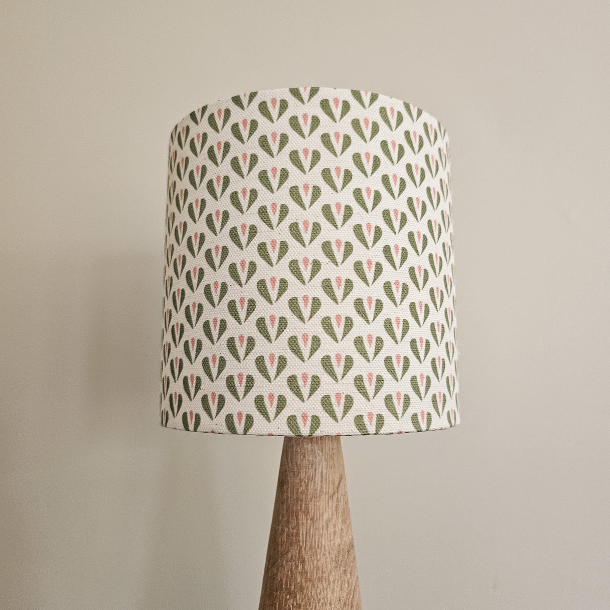A table lamp with a drum shade featuring a repeating green and pink heart pattern on a white background, supported by a tapered natural wood base against a plain beige wall.
