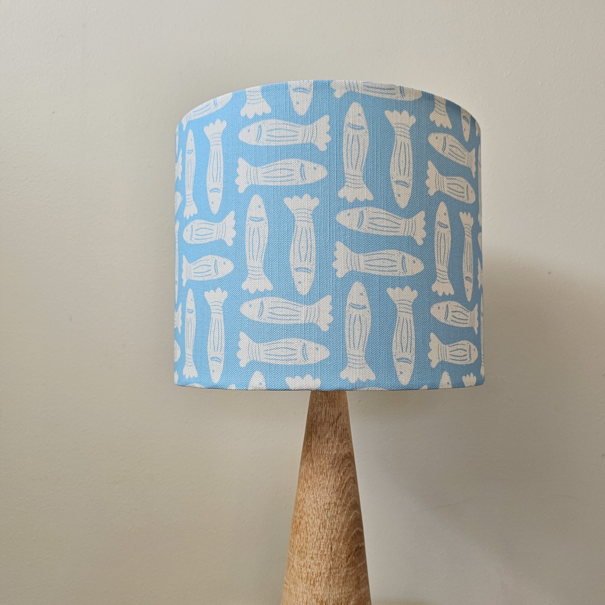 Zia Living Fishes fabric swatch lino print in sky blue with white fish featured on a drum lampshade on a lamp base