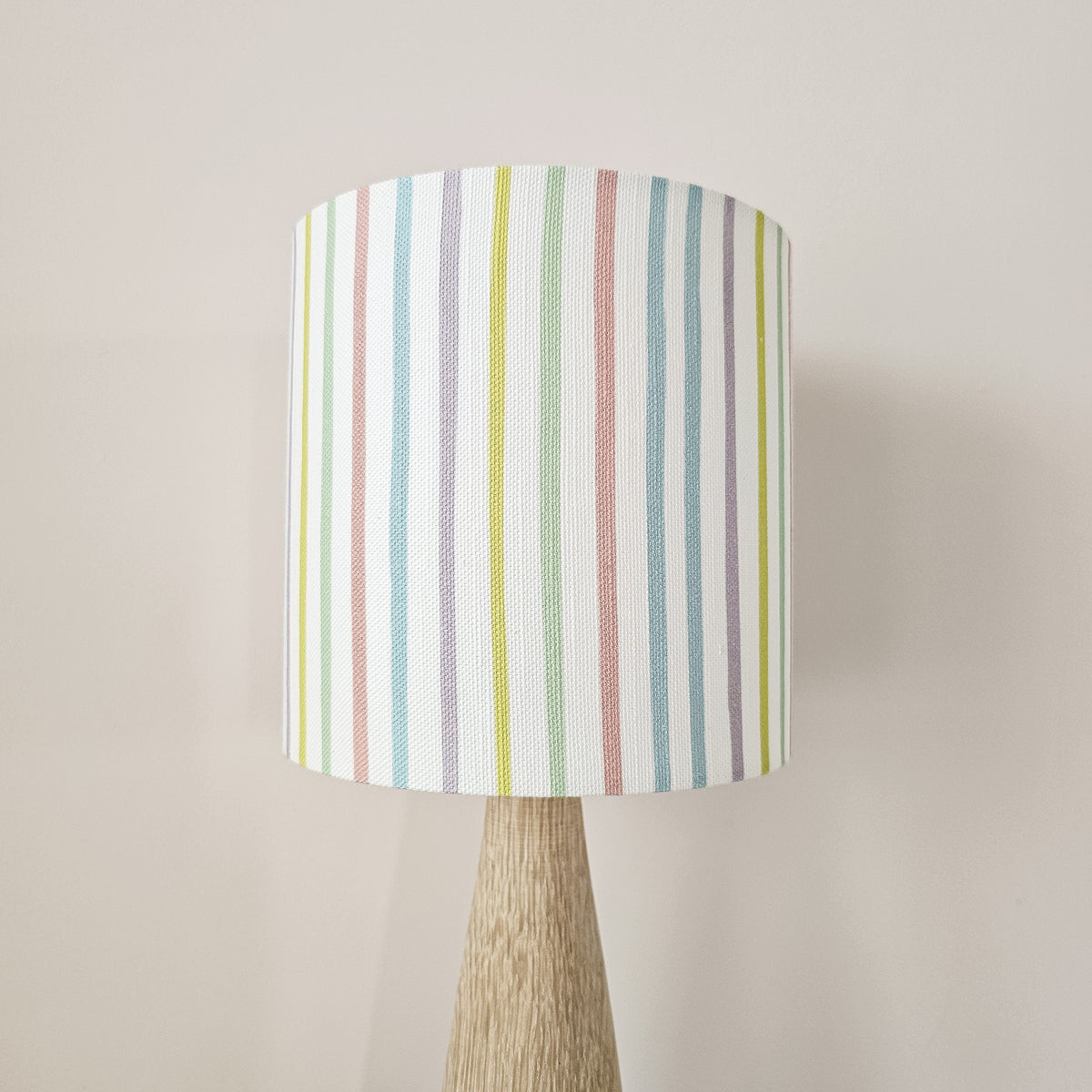 A table lamp with a white drum shade featuring vertical pastel stripes in blue, green, pink, and yellow, mounted on a tapered light wood base against a plain beige wall.