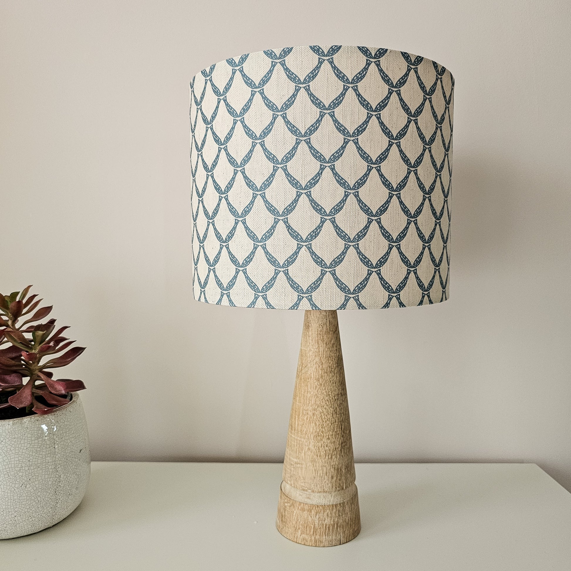 Marina cotton linen fabric featuring a repeating blue curved geometric trellis motif on an off‑white textured background. Featured on a lampshade and wooden base on side board with flower ZIA LIVING.