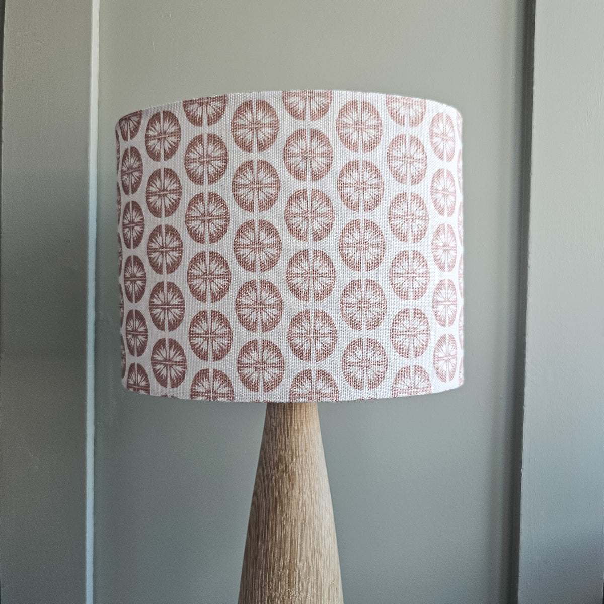 A table lamp with a tapered natural wood base and a drum shade featuring a repeating dusty rose circular pressed flower pattern on a white fabric background, set against a green panelled wall.