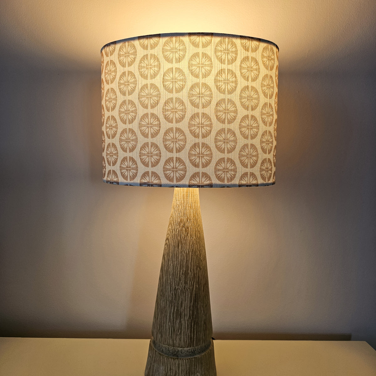 A table lamp with a tapered natural wood base and a drum shade featuring a repeating dusty rose circular pressed flower pattern on a white fabric background, with lamp on with cosy glow