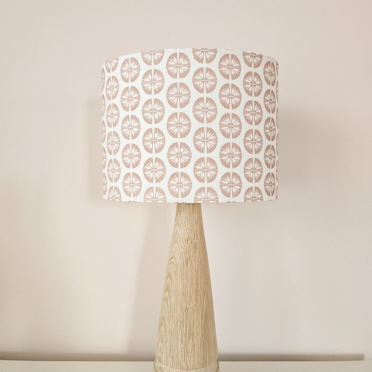 A table lamp with a tapered natural wood base and a drum shade featuring a repeating dusty rose circular pressed flower pattern on a white fabric background, set against a pale pink background