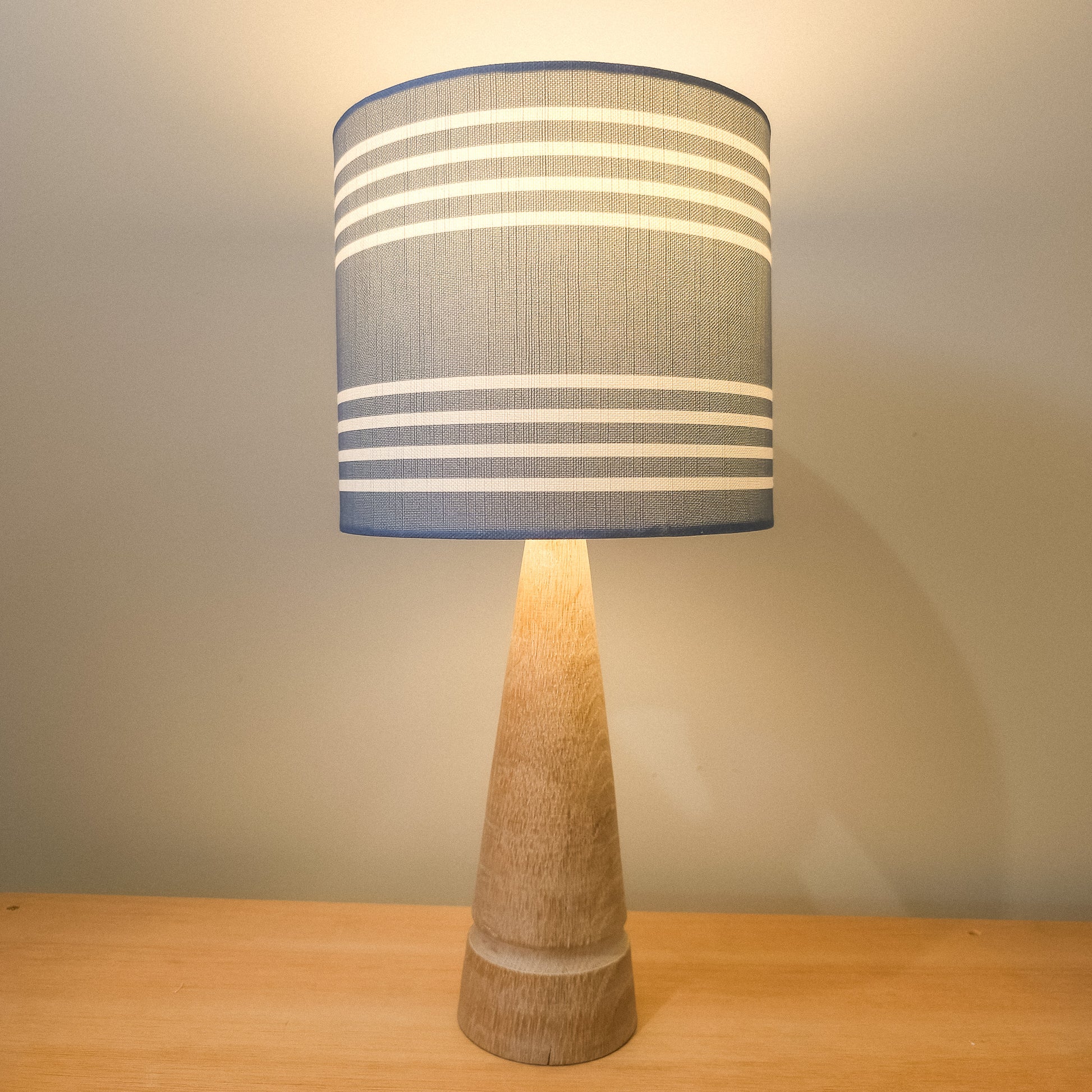Drum lampshade in Blue Studio Stripe fabric on a wooden lamp base, softly lit to show the horizontal stripe pattern — artisan home décor by ZIA LIVING.