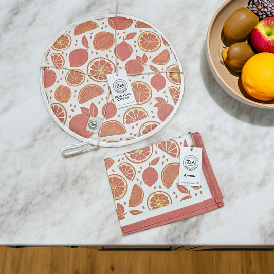 A round Moro Orange aga hob cover and matching tea towel from ZIA LIVING arranged on a marble surface beside a wooden bowl of fruit.